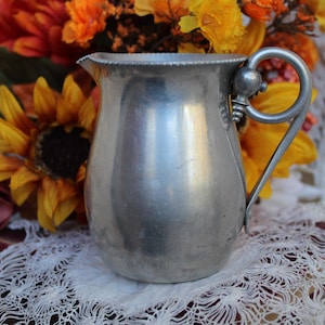 Könnte beinhalten: Eine silberne Kanne mit rundem Körper und einem dekorativen Henkel. Die Kanne steht auf einer weißen Spitzentischdecke.