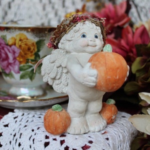 May include: A white cherub figurine with wings holding an orange pumpkin. The cherub is wearing a brown wreath and has blue eyes. The figurine is sitting on a white crocheted surface with two smaller pumpkins.