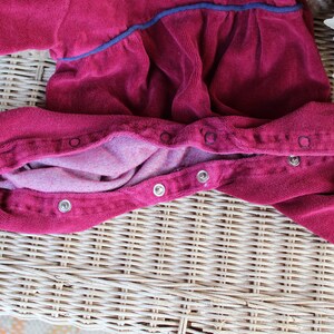 May include: A red corduroy baby onesie with a snap closure down the front. The onesie is laid out flat on a wicker surface.