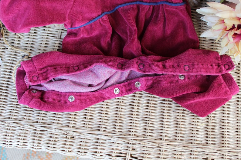 May include: A red corduroy baby onesie with a snap closure down the front. The onesie is laid out flat on a wicker surface.