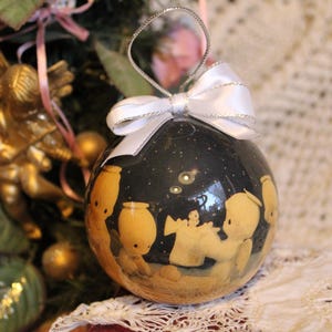 May include: A spherical Christmas bauble with a dark blue backdrop and a painted scene of figures. The bauble has a silver loop and a white bow. The bauble is hanging on a Christmas tree.