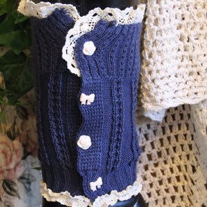 Op de afbeelding: Blauwe gebreide beenwarmers met witte kantversiering en witte knoopaccenten. De beenwarmers zijn op een mannequin en worden getoond tegen een witte gehaakte achtergrond.