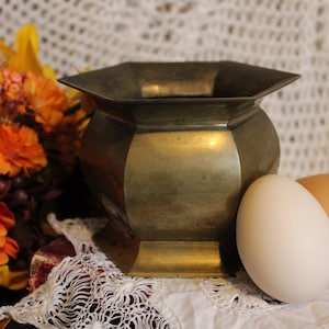 May include: A gold-colored metal planter with a hexagonal shape and a smooth, shiny surface. The planter is sitting on a white crocheted doily with two eggs beside it.