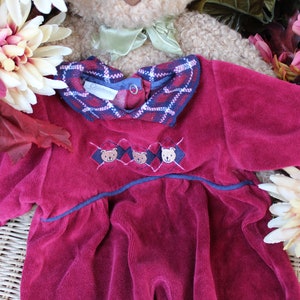 May include: A burgundy velvet baby dress with a plaid collar and embroidered bears. The dress has a navy blue trim and a ruffled hem.
