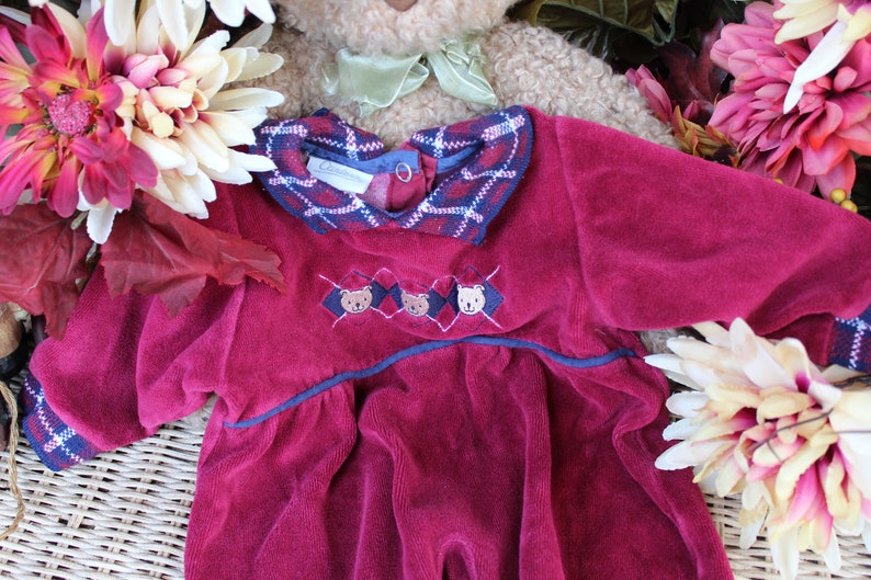 May include: A burgundy velvet baby dress with a plaid collar and embroidered bears. The dress has a navy blue trim and a ruffled hem.
