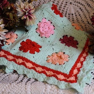 Könnte beinhalten: Eine gehäkelte Babydecke mit hellblauem Hintergrund und bunten Blumendesigns. Die Decke hat einen roten und rosafarbenen Rand.