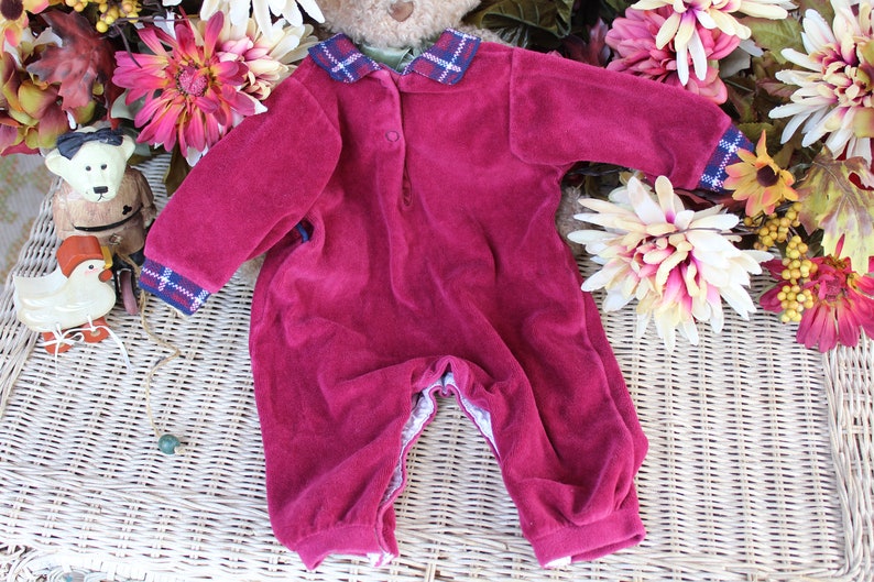 May include: A burgundy velour baby onesie with a plaid collar and cuffs. The onesie has a button closure at the neck and a snap closure at the crotch.