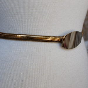 Może przedstawiać: Rozciągliwy pasek w kolorze złota z teksturowanym, żebrowanym wzorem. Pasek posiada owalne, marmurkowe zapięcie w odcieniach brązu i beżu. Pasek jest prezentowany na białym manekinie, prezentując jego styl i design.