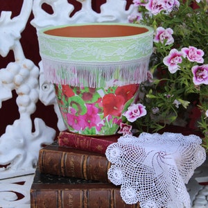 May include: A terracotta flower pot with a green and white lace trim and a pink and green floral pattern. The pot is sitting on a stack of old books with a white crocheted doily on top.