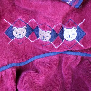 May include: A burgundy velvet fabric with a blue trim and embroidered teddy bear designs. The bears are in a diamond pattern with white stitching.