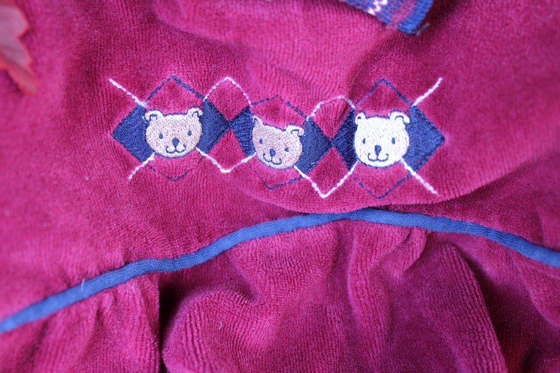May include: A burgundy velvet fabric with a blue trim and embroidered teddy bear designs. The bears are in a diamond pattern with white stitching.