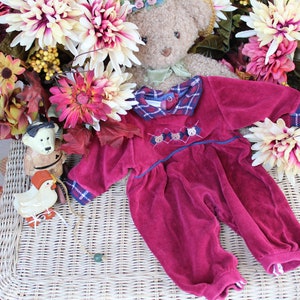 May include: A plush burgundy baby onesie with a plaid collar and embroidered bear faces. The onesie is lying on a wicker surface with a teddy bear and fall-themed flowers in the background.