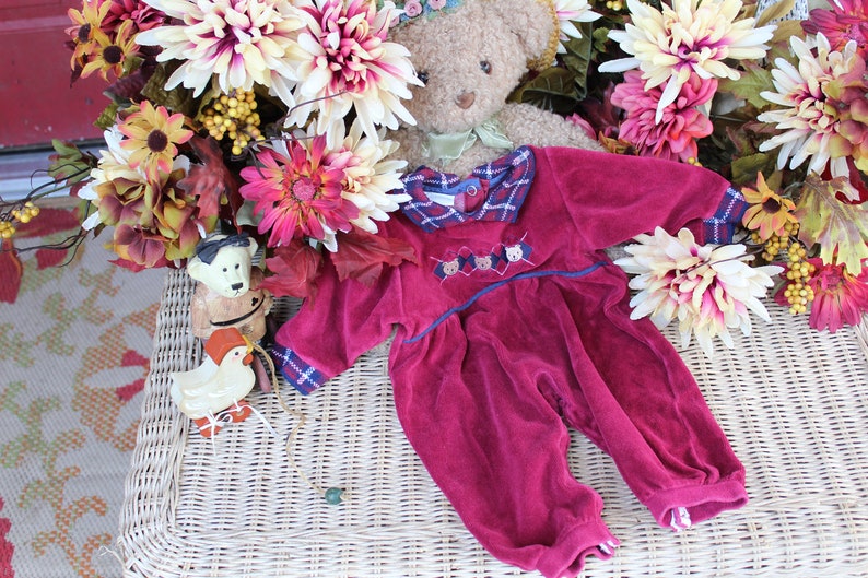 May include: A plush burgundy baby onesie with a plaid collar and embroidered bear faces. The onesie is lying on a wicker surface with a teddy bear and fall-themed flowers in the background.