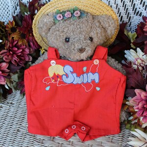 Puede incluir: Un oso de peluche con un sombrero de paja con un adorno floral y un pelele rojo. El pelele tiene la palabra "Swim" bordada, junto con un dibujo animado. El fondo presenta flores coloridas.