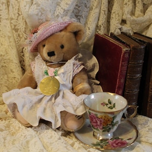 May include: A brown teddy bear in a pink hat and white dress, with a gold medallion, sits near antique books and a floral teacup and saucer. The scene is set against a backdrop of lace.