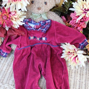 May include: A burgundy velour baby onesie with a plaid collar and embroidered teddy bears. The onesie is laid out on a wicker surface with a teddy bear wearing a straw hat in the background.