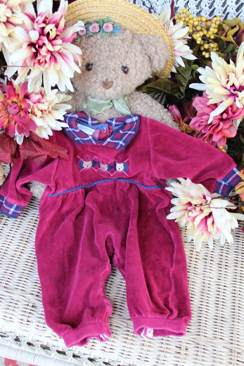 May include: A burgundy velour baby onesie with a plaid collar and embroidered teddy bears. The onesie is laid out on a wicker surface with a teddy bear wearing a straw hat in the background.
