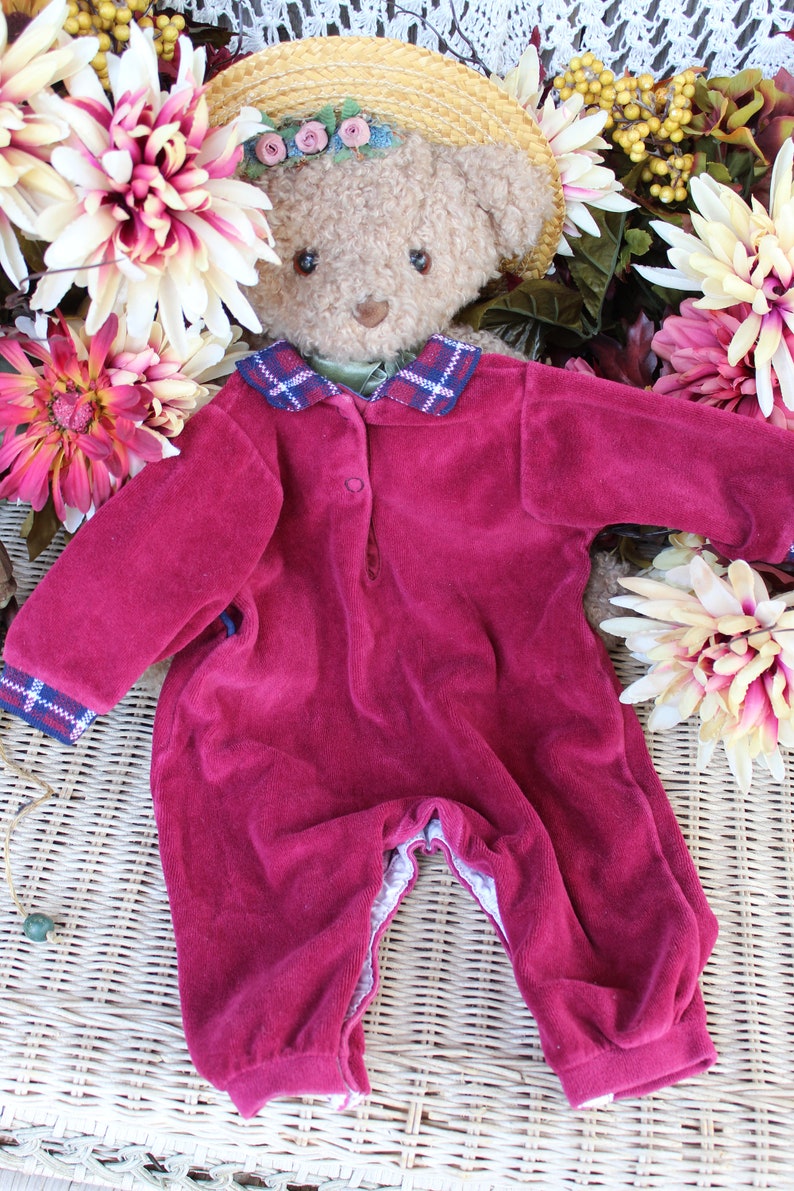 May include: A plush teddy bear wearing a burgundy velvet jumpsuit with a plaid collar and a straw hat with floral embellishments.
