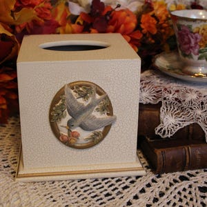 May include: White ceramic tissue box cover with a raised design of a blue bird in a gold frame. The bird is surrounded by leaves and berries.