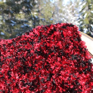 Pode incluir: Um close-up de um cachecol fofo e texturizado em uma mistura de vermelho e preto. O cachecol parece ser feito de um material macio e felpudo. O fundo está desfocado, com uma paisagem nevada e árvores visíveis.