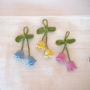 Könnte beinhalten: Drei gehäkelte Blumenanhänger mit grünen Blättern und glockenförmigen Blüten in Blau, Gelb und Rosa.