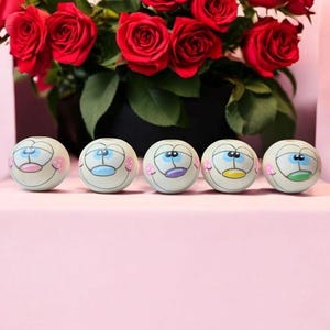 Könnte beinhalten: Fünf weiße Keramik-Kugeln mit verschiedenen farbigen Gesichtern. Jede Kugel hat ein blaues Auge und einen andersfarbigen Mund. Die Münder sind rosa, blau, lila, gelb und grün.