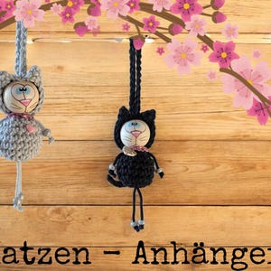 May include: Two handmade crochet cat ornaments, one grey and one black, hang against a wooden background. Each cat has a painted face, a pink heart, and a decorative collar. The background features pink and purple flower blossoms.