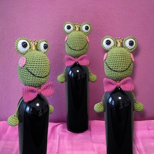Könnte beinhalten: Drei gehäkelte Frosch-Flaschenverschlüsse mit rosa Schleifen und goldenen Kronen. Die Frösche sind grün und haben schwarze Augen. Die Flaschenverschlüsse befinden sich auf Weinflaschen.