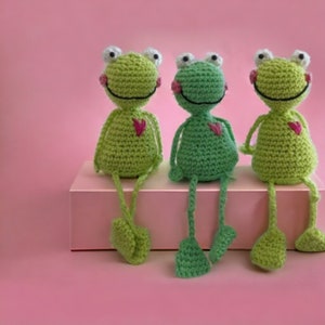 Könnte beinhalten: Drei gehäkelte grüne Frosch-Plüschtiere mit rosa Herzen auf der Brust sitzen auf einer rosa Oberfläche. Sie haben weiße Augen und schwarze Lächeln.
