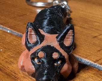 May include: A black and brown 3D printed German Shepherd dog keychain. The dog is in a lying down position with its head facing forward. The keychain has a silver metal ring.