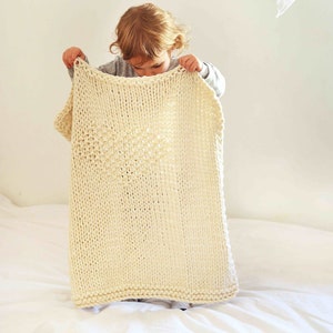 May include: A child holds a large, cream-colored knitted blanket with a heart-shaped pattern. The blanket is made of thick yarn and has a soft, fluffy texture.