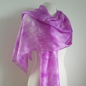 May include: A lilac-coloured silk scarf with a tie-dye pattern, displayed on a mannequin. The scarf's material is semi-sheer, featuring white highlights. This scarf is a fashion accessory.