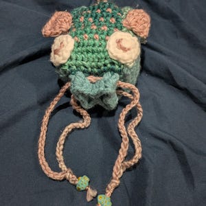 May include: A crocheted green and pink creature with large eyes and a long pink cord. The cord has two blue and gold fish-shaped beads attached to it.