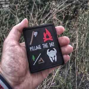 Peut inclure: Un écusson brodé noir avec une hache blanche, un feu de camp rouge, un arbre vert, une tente brune et un casque blanc avec une visière noire. Le texte "Pillage The Day" est brodé en blanc sur l'écusson.
