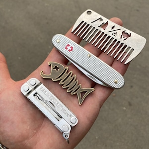May include: A silver pocket knife with a red cross logo, a silver multi-tool with the text "LEATHERMAN Micra", and a silver comb with a cut-out design of a mountain, a tree, and a buffalo.