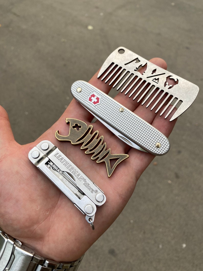 May include: A silver pocket knife with a red cross logo, a silver multi-tool with the text "LEATHERMAN Micra", and a silver comb with a cut-out design of a mountain, a tree, and a buffalo.