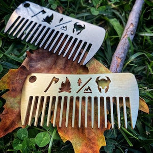 May include: Two silver metal pocket combs with a camping theme. The combs have a variety of symbols including an axe, a campfire, a mountain, and a viking helmet.