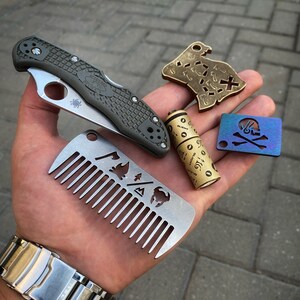May include: A hand holding a variety of small metal objects, including a pocket knife, a comb, a small metal tube, a brass map-shaped keychain, and a blue skull and crossbones keychain.