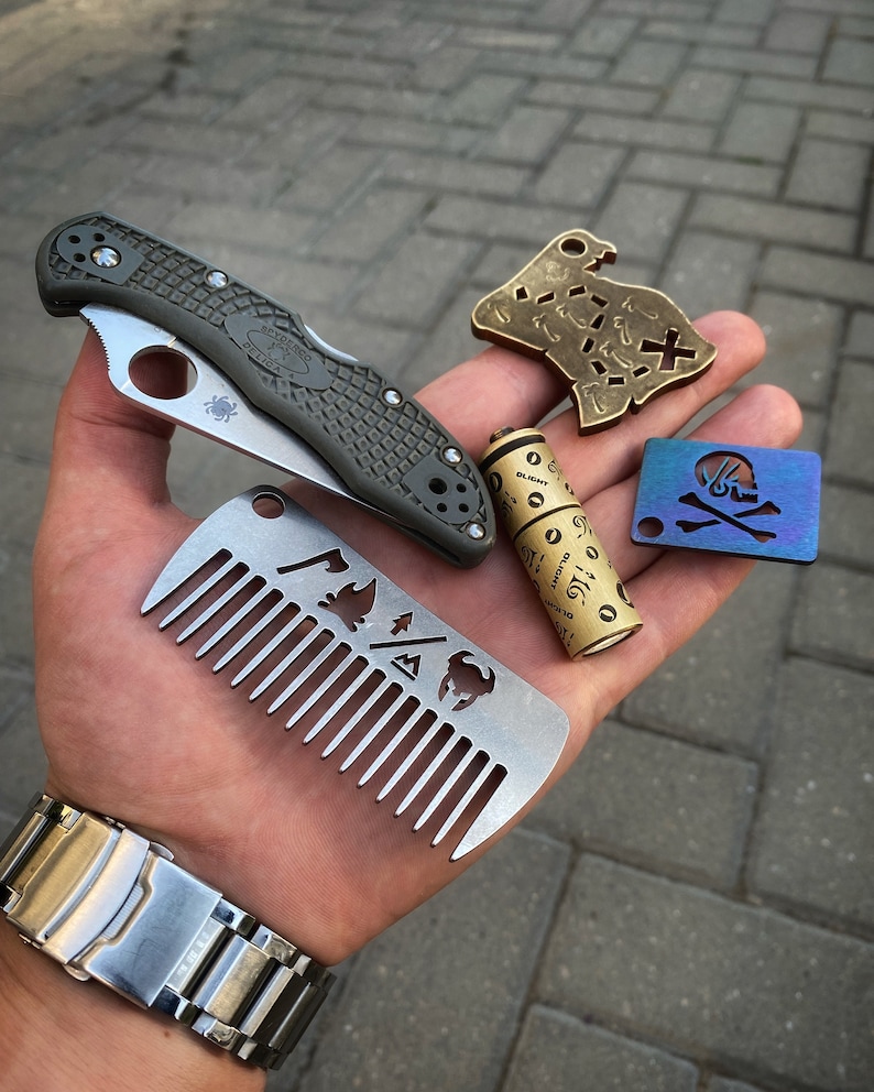 May include: A hand holding a variety of small metal objects, including a pocket knife, a comb, a small metal tube, a brass map-shaped keychain, and a blue skull and crossbones keychain.