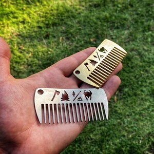May include: Two metal pocket combs with different designs. The top comb is gold and features a campfire, axe, and tree. The bottom comb is silver and features a campfire, axe, tree, and a mountain.