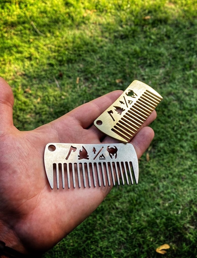 May include: Two metal pocket combs with different designs. The top comb is gold and features a campfire, axe, and tree. The bottom comb is silver and features a campfire, axe, tree, and a mountain.
