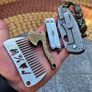 May include: A set of four pocket-sized tools, including a comb, a bottle opener, a multi-tool, and a folding knife. The tools are made of metal and have a silver finish. The comb has a fire, a tree, and an axe design. The bottle opener has a skull and crossbones design. The multi-tool has the text "Leatherman Micra" on it. The folding knife has a green and brown paracord lanyard.