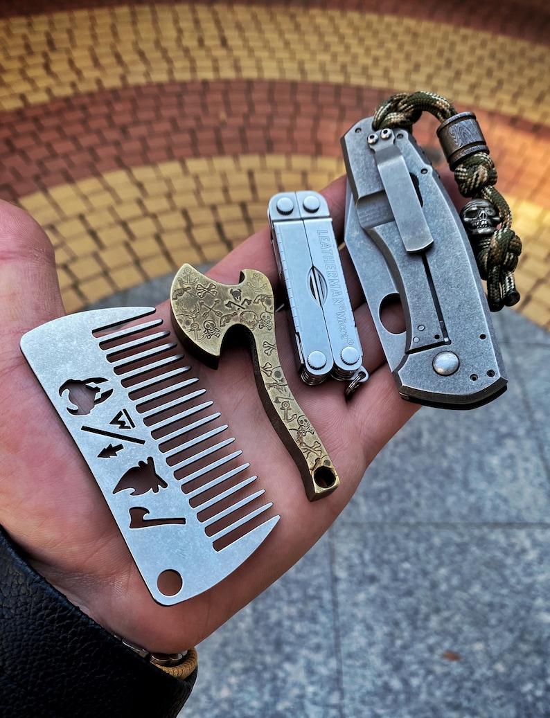 May include: A set of four pocket-sized tools, including a comb, a bottle opener, a multi-tool, and a folding knife. The tools are made of metal and have a silver finish. The comb has a fire, a tree, and an axe design. The bottle opener has a skull and crossbones design. The multi-tool has the text "Leatherman Micra" on it. The folding knife has a green and brown paracord lanyard.
