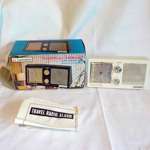 May include: A vintage Windsor Solid State AM Radio alarm clock. The clock is white with a silver clock face and a speaker grill. The original box is teal and black with the words "Windsor" and "Solid State AM Radio" printed on it. A paper reads "Travel Radio Alarm".