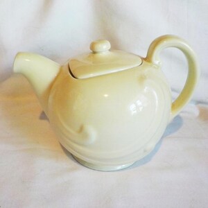May include: A pale yellow ceramic teapot featuring a rounded body with embossed swirling scroll patterns, a curved handle, a short spout, and a matching lid with a small knob handle, presented against a plain white background.