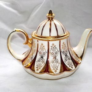 Op de afbeelding: Een vintage theepot met een crèmekleurige basis, gouden accenten en een bordeauxrode band. Het deksel is bekroond met een gouden knop. De theepot heeft een gebogen handvat en tuit, met een sierlijk ontwerp.