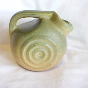May include: A small, handmade ceramic pitcher in shades of green and tan. The pitcher has a rounded body with a handle and a spout. The base features a spiral design. This decorative piece is suitable for serving beverages.