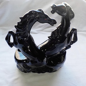 Könnte beinhalten: Schwarze Keramikskulptur von zwei sich aufbäumenden Pferden, die sich gegenüberstehen. Die Pferde haben rote Augen und eine glänzende Oberfläche. Die Skulptur ist ein Dekorationsartikel.