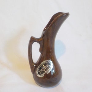 Könnte beinhalten: Ein kleiner, brauner Keramikkrug mit gebogenem Ausguss und Henkel. Der Krug hat eine glänzende Oberfläche und ein Etikett mit der Aufschrift "DriDen". Das Design ist schlicht und elegant, geeignet zum Servieren von Saucen oder kleinen Portionen.