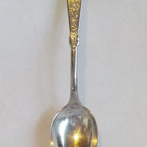 May include: A silver-toned metal spoon featuring an elongated handle with an embossed relief design of a small figure and floral motifs. The spoon has a smooth, oval-shaped bowl and a polished finish that reflects light across its surface.